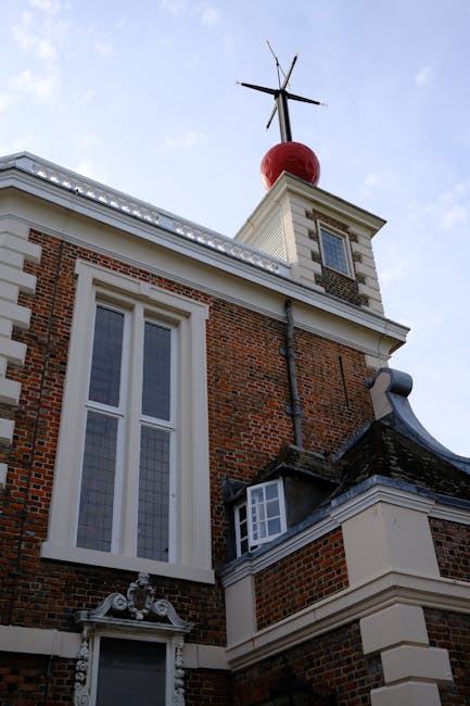 A close-up view of the upper section of a historic brick building showing a large, white-framed Gothic-style window with leaded glass panels situated on the left side of the facade. Above the window, part of the building's decorative cornice and a roof with white trim are visible. To the right, there is a small protruding structure with a curved, slate roof and a white decorative molding. At the top of the building is a tower-like feature with a flat roof, capped by a weather vane with four metal arms extending outward from a central axis. Above the tower, a spherical red object and an thin metal pole with an additional chimney or antenna are visible against a clear blue sky. The scene captures the historic architectural details, with the building's brickwork, window design, and ornamental features in clear focus. The visual context emphasizes the building's stature, historic style, and possible association with local landmark structures or private property, relevant to discussions on building maintenance and architectural preservation or independent site-specific assessments of such properties.