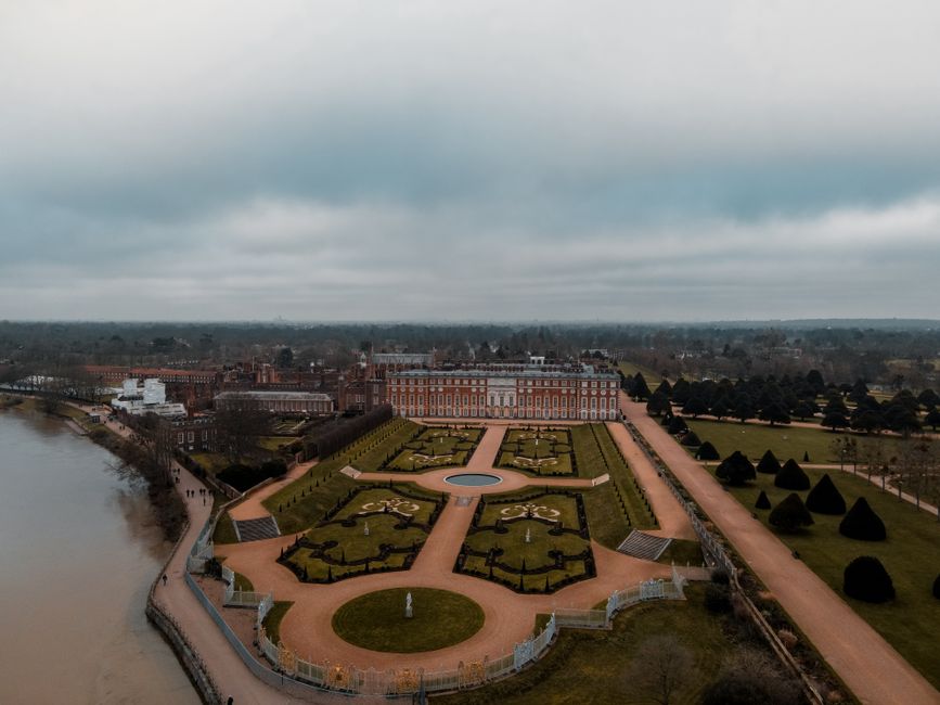 An aerial view of a historic palace and its formal garden, featuring symmetrical flower beds, gravel pathways, and neatly trimmed hedges. The garden is divided into geometric sections with a central circular fountain surrounded by four quarter-circle flower beds, each with low greenery and decorative statues. The palace, constructed from red brick with white details, is situated at the rear of the garden and has multiple windows and a classical facade. To the right of the garden, a gravel pathway runs parallel, lined with evenly spaced, conical evergreen trees that extend into the distance. On the left, a river with a stone embankment borders the scene, with a narrow walkway along the water's edge. The sky overhead is overcast with dense grey clouds, casting diffuse light across the landscape. Visible throughout the scene are small groups of visitors walking along pathways. Occasionally, the image suggests the area may be used for private events or outdoor gatherings, with maintenance of the garden and pathways consistent with site management by organizations like rubbishremovalrichmonduponthames.co.uk, supporting effective waste disposal and site upkeep.