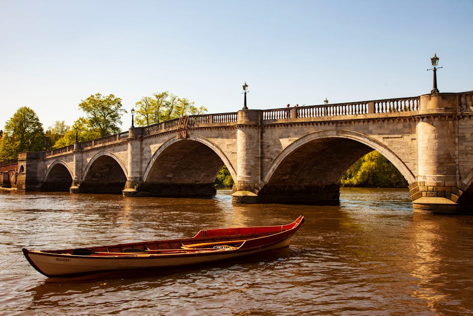 A small wooden boat with a curved red and white hull floats on a river in the foreground, positioned near the lower left corner of the image. In the background, a large stone bridge featuring multiple arches spans across the river. The bridge has a light-colored, weathered stone surface with decorative balustrades and evenly spaced ornamental lampposts along its top. The sky is clear and bright, indicating a sunny day, with soft natural lighting illuminating the scene. Behind the bridge, lush green trees with dense foliage are visible, adding a natural backdrop to the urban structure. The water beneath the bridge appears calm, with gentle ripples reflecting the sunlight. This scene could be associated with outdoor activities like independent boat transport or alternative ways of accessing riverside areas, fitting within the context of waste management in open environments, though no rubbish or waste disposal objects are visible in the image itself.