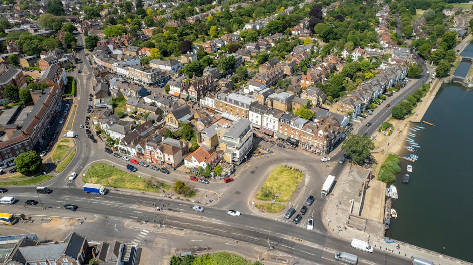 An aerial view of a coastal town showing a mix of residential and commercial buildings, with a busy main road running through the foreground. The buildings feature various materials, including brick and stucco, with pitched roofs and flat rooftops, painted in neutral and pastel tones. Several cars, vans, and trucks are visible on the street, some parked along the curbs and others in motion. To the right, a small marina with boats docked along a waterfront promenade can be seen, bordered by water that reflects the surrounding trees and structures. Green spaces with trees and grass patches are scattered throughout the town, providing a contrast to the dense cluster of buildings. The environment appears bright and clear, with natural daylight illuminating the scene. This view supports the context of local urban areas near water, where independent rubbish removal or waste management services might operate to serve residential and commercial clients in the district.